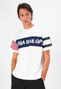 White short-sleeve t-shirt featuring a navy blue "USA SAIL GP" graphic across the chest, striped red and white sleeve, and black accents.