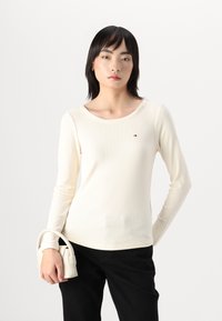 Langärmliges, cremefarbenes Rippoberteil mit rundem Ausschnitt und kleinem Logo. Kombiniert mit schwarzen Hosen und einer kleinen weißen Handtasche.