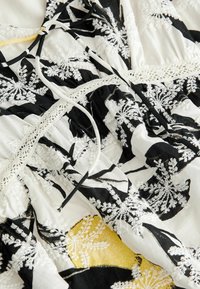 Tissu brodé noir et blanc avec des motifs floraux ; présente des accents en dentelle et une section jaune, stylé avec des liens réglables.