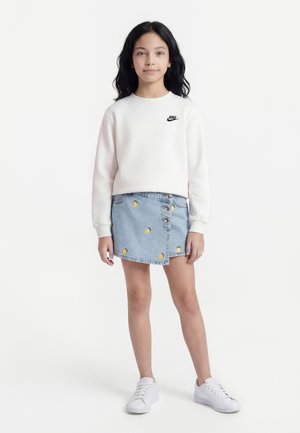 Fată tânără în picioare, purtând un hanorac alb Nike, fustă de denim albastru deschis cu modele de lămâie și adidași albi, pe un fundal alb.