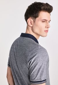 Szary polo z granatowym kołnierzykiem i paskowanymi mankietami. Posiada teksturowany materiał i dopasowany krój, widoczny z lekko obróconego profilu.