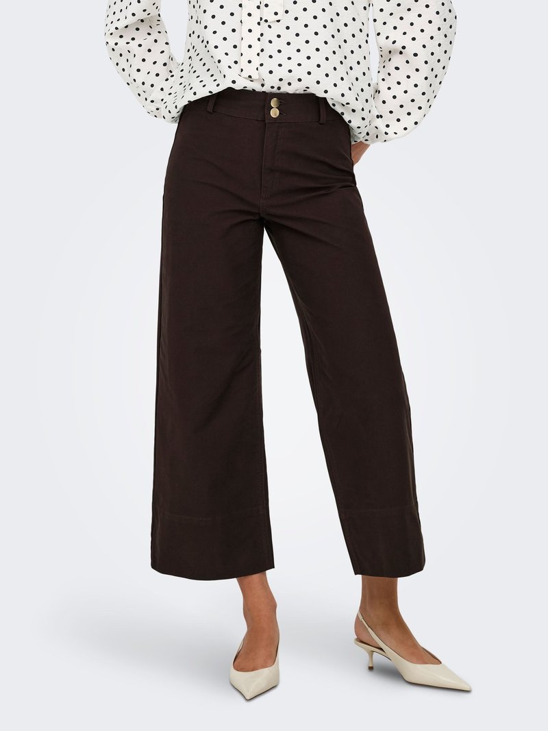 Femme portant un pantalon marron foncé taille haute à jambes larges avec un devant croisé boutonné et des escarpins slingback blancs à bout pointu.