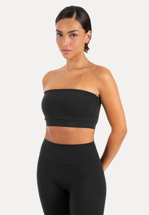 Kvinde med kort mørkt hår iført en sort stropløs cropped top og høj taljede sorte leggings mod en hvid baggrund.