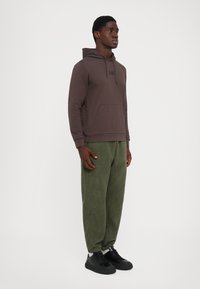 Ung mann står iført brun hettegenser, grønne joggebukser, hvite sokker og svarte joggesko på hvit bakgrunn.
