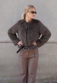 Jachetă scurtă din blană ecologică maro, cu mâneci lungi, închidere cu fermoar și curea neagră din piele. Asortată cu pantaloni maro cu picioare largi.