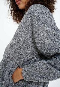 Kobieta z kręconymi włosami, ubrana w szary wełniany płaszcz o teksturowanej powierzchni, z jedną ręką w kieszeni, na białym tle.