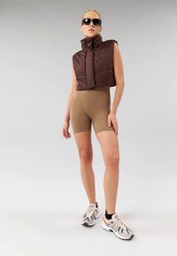 Pruun lühike puffervest, beež kõrge vöökohaga lühikesed püksid ja heledad tossud mitmevärviliste aktsentidega. Modell kannab musti päikeseprille; taust on lihtne.