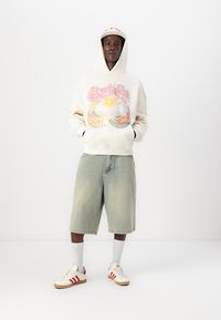 Cream hoodie with graphic print, light blue denim shorts, white socks, and cream sneakers with red stripes. Short sleeves; relaxed fit.