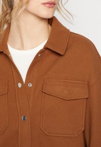 Brun, texturerad, ullaktig skjorta med jacka som har en krage, tryckknappar och två bröstfickor. Underlag är en vit topp.