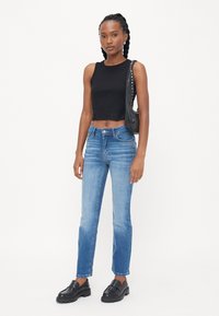 Svart avkortet tanktopp, lyseblå straight-leg jeans og svarte slip-on sko. Modell som holder en svart bag med nagler, står mot en hvit bakgrunn.