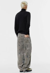 Oseba s kratkimi blond lasmi nosi črn pulover z visokim ovratnikom in prevelike sive hlače s madeži, stoji z rokami v žepih na hrbtni strani proti belemu ozadju.