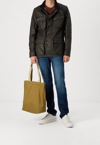 Tmavě zelená voskovaná bunda s několika kapsami, hnědý kordový límec, modré džíny, béžová plátěná taška a bílé tenisky.