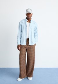 Mand iført lyseblå skjorte med knapper, hvid tanktop, brune ternede bukser med vid ben, hvide sneakers, grå kasket og perlesnor står på blå gulv.