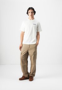 Cream cotton t-shirt with Timberland logo, beige cargo pants with side pockets, and brown leather boat shoes. Simple, casual outfit.