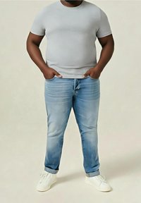Homme debout les mains dans les poches, portant un t-shirt gris clair, un jean bleu avec les revers roulés et des baskets blanches sur un fond uni.