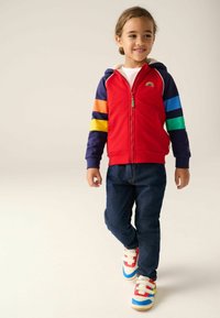 Felpa rossa con zip, strisce arcobaleno, maniche blu e un piccolo dettaglio arcobaleno. Abbinata a jeans blu e scarpe sportive multicolori.
