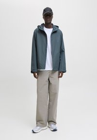 Person iført en grå hættejakke, hvid t-shirt, beige bukser med vide ben, sølvfarvede sneakers og sort kasket, der står på en ensfarvet hvid baggrund.