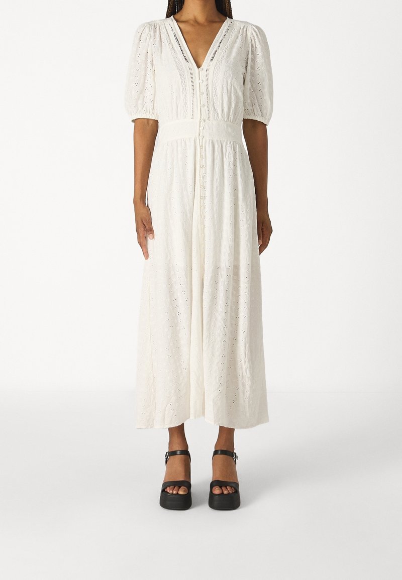 Femme portant une robe midi blanche en œillet avec des manches bouffantes et un devant à boutons, associée à des sandales à plateforme noires.