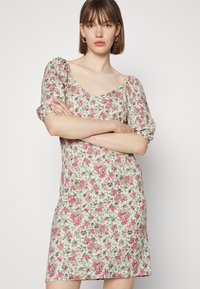 Vestido floral com um fundo claro e flores rosas e vermelhas. Possui um decote redondo e mangas curtas bufantes, feito de um tecido suave.