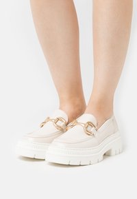 Krémové loafers s robustnou bielou podrážkou, s pozláteným reťazovým akcentom cez vrch. Hladký kožený materiál a minimalistický dizajn.