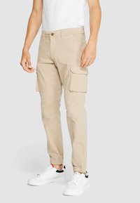 Beige Cargo-Hosen mit schmal zulaufendem Bein, seitlichen Cargo-Taschen und elastischen Bündchen, aus einem Baumwollmischgewebe, getragen mit weißen Sneakers.