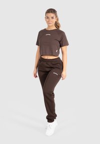 Brun cropped t-shirt med hvidt logo og matchende brune joggers, der har elastisk talje og manchetter ved anklerne, parret med hvide sneakers.