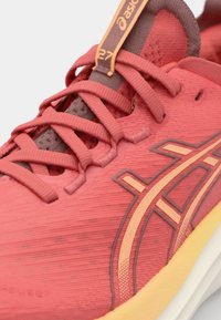 Chaussure de course rouge avec un dessus texturé, ornée de lacets et d'accents marron. Elle possède un logo distinctif et une semelle rembourrée.