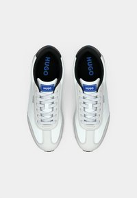 Paire de baskets blanches Hugo avec lacets gris, logo bleu sur la languette et la semelle intérieure, vues de dessus sur fond blanc.