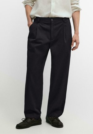 Uomo con pantaloni neri plissettati, scarpe nere e camicia beige a maniche lunghe, mani rilassate ai lati, in piedi su sfondo semplice.