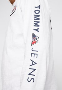 Vit långärmad skjorta med marinblå text "TOMMY" och "JEANS" på ärmen, samt en logotyp på framsidan med en triangulär design.