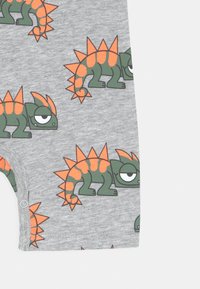 Šedý bavlněný overal s opakujícím se vzorem dinosaurů, se zelenými těly a oranžovými hřebeny. Zapínání na patentky na spodní části.