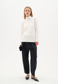 Maglione bianco a collo alto con orlo a costine, abbinato a pantaloni neri larghi e ballerine nere. La modella tiene una piccola borsa a tracolla nera.