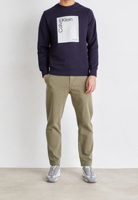 Marinblå sweatshirt med en vit rektangulär logotyp, ihop med ljusgröna tapered-byxor och grå sneakers. Slät tyg och avslappnad design.