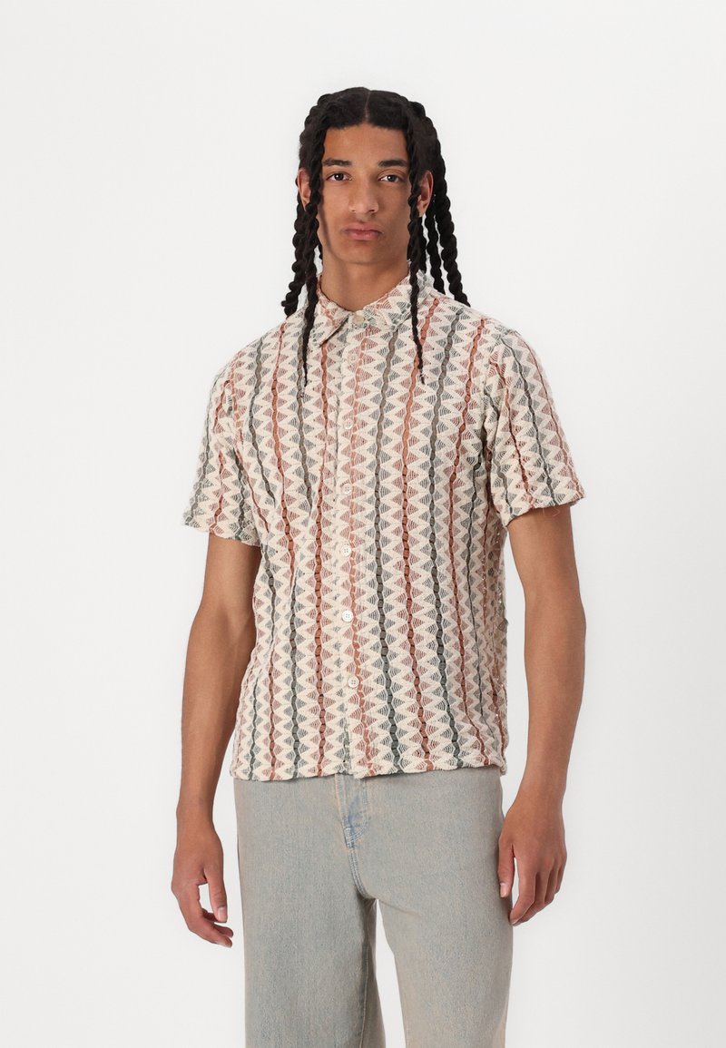 Camisa de manga corta con botones que presenta una base color crema con patrones geométricos repetitivos en rojo, verde y gris. Tejido texturizado.