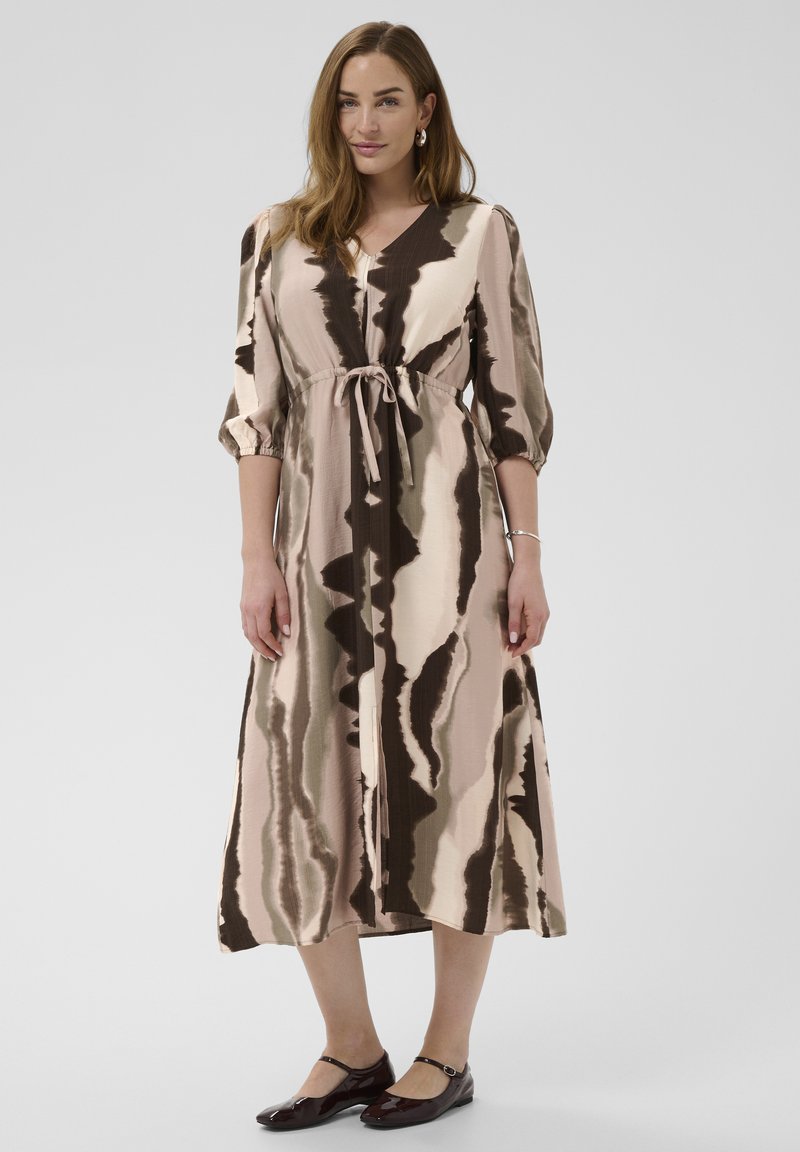 Donna che indossa un abito midi tie-dye beige e marrone con maniche a sbuffo 3/4, vita annodata e ballerine Mary Jane scure, in piedi su uno sfondo semplice.