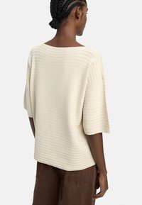 Jasno beżowy sweter prążkowany z szerokim dekoltem i krótkimi rękawami. Teksturowana tkanina ma pionowe linie, zestawiona z brązowymi spodniami.