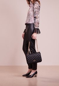 Mujer con blusa de estampado y puños de encaje, pantalones de cuero negros, tacones negros de bloque, sosteniendo un bolso acolchado negro con cadena dorada.