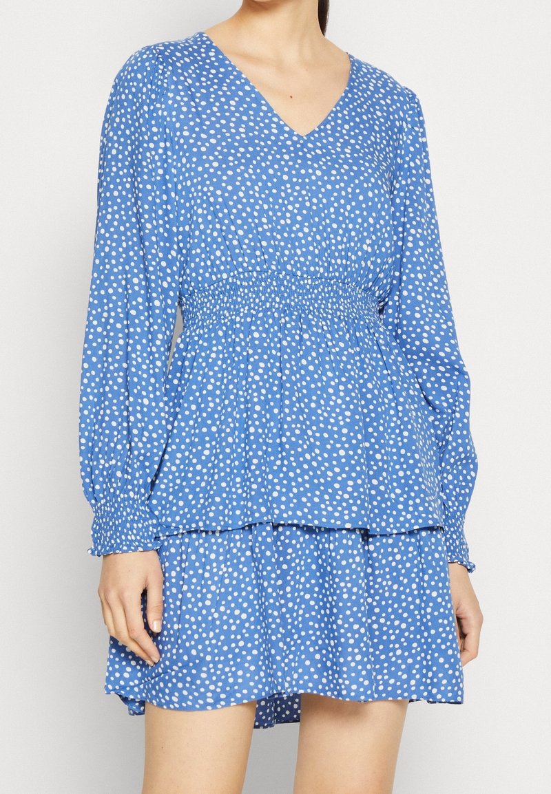 Robe bleue avec un col en V, des manches longues et des pois blancs. Elle comporte une taille smockée et une jupe évasée à volants en couches.