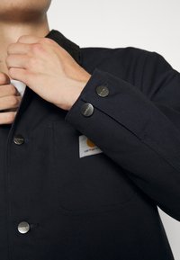 Marineblaues Jacket mit einem Cordkragen, ausgestattet mit metallischen Druckknöpfen und einer Brusttasche mit einem Logopatch. Strukturierter Stoff ist sichtbar.