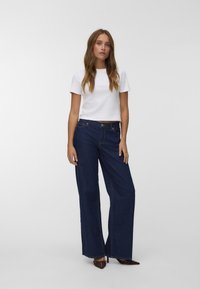Bela skrajšana majica in temno modri široki jeans z detajli šivanja. Model stoji na nevtralni podlagi, oblečen v špičaste čevlje.
