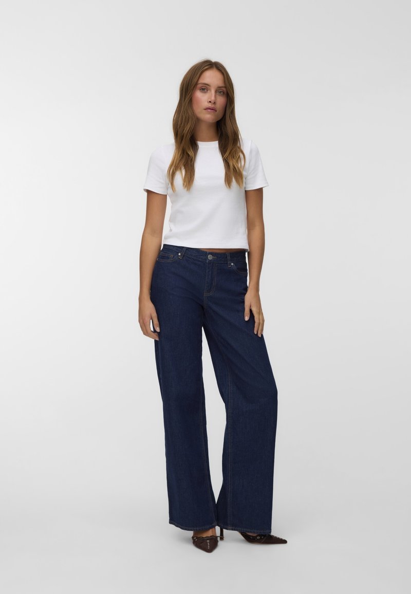 Bela skrajšana majica in temno modri široki jeans z detajli šivanja. Model stoji na nevtralni podlagi, oblečen v špičaste čevlje.