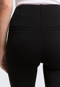 Hoch taillierte schwarze Hose aus glattem Stoff mit zwei Gesäßtaschen und schmaler Passform, die eine nahtlose Verarbeitung und klare Linien zeigt.