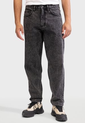 Personne portant un jean noir effet délavé à l'acide et des baskets épaisses beige et noires, debout devant un fond neutre.