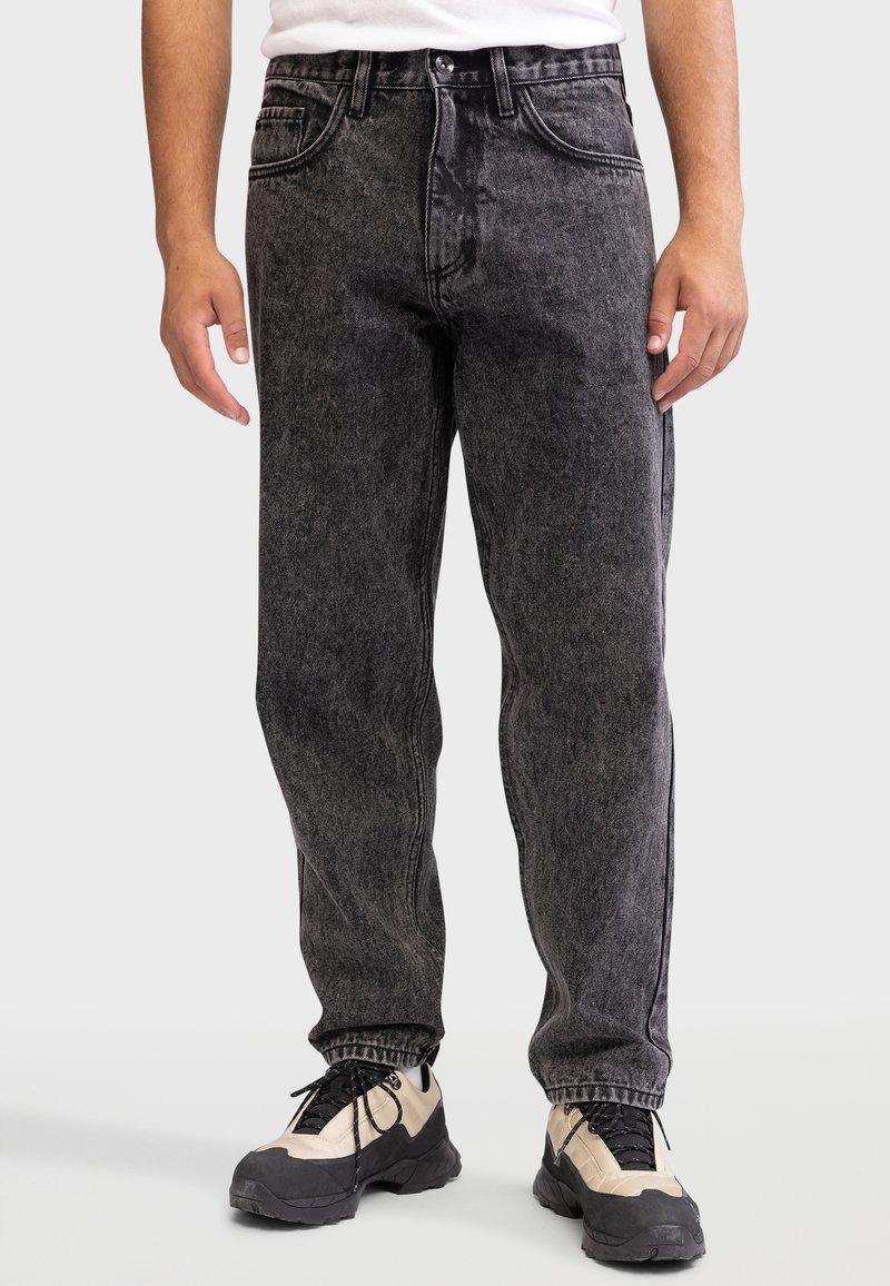Personne portant un jean noir effet délavé à l'acide et des baskets épaisses beige et noires, debout devant un fond neutre.