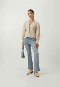 Cardigan beige in maglia con motivo texturizzato, chiusura a bottoni, abbinato a jeans svasati azzurri chiari e stivali con tacco beige, mentre tiene una piccola borsa blu.