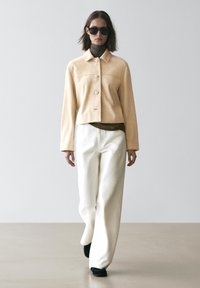 Beige croppede ruskindjakke med to store knapper, båret over en mørk rullekrave og hvide bukser med vide ben, kombineret med sorte sko.