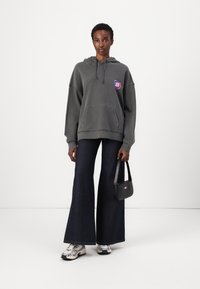 Sweatshirt gris à capuche avec poche avant, arborant un écusson de logo coloré, associé à un jean flare noir et des baskets argentées. Sac à main noir.