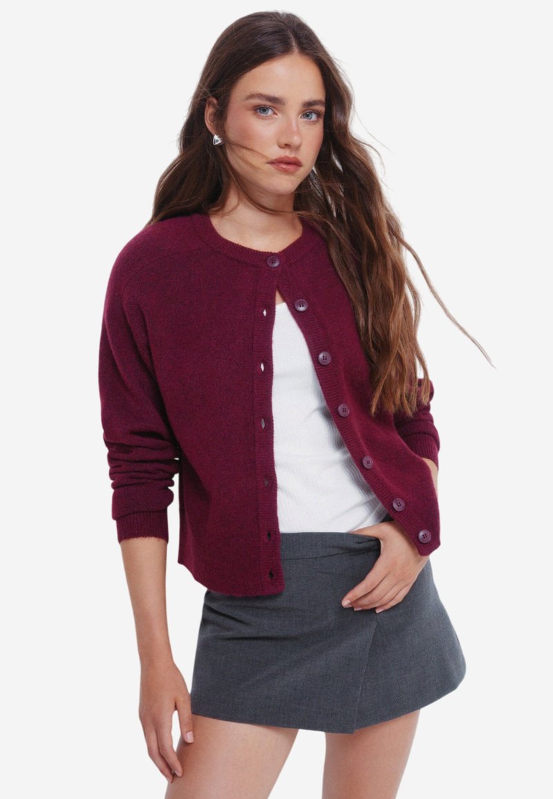Burgundský cardigan s kulatým výstřihem, zapínáním na knoflíky vpředu, žebrovanými manžetami a měkkou texturou, v kombinaci s přiléhavou šedou sukni.