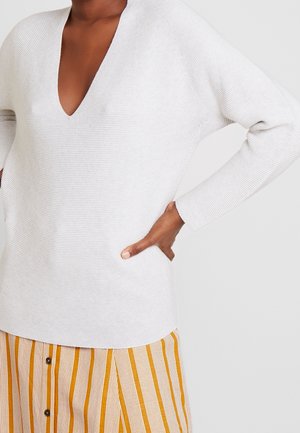 Maglione grigio chiaro lavorato a maglia con scollatura a V profonda e maniche lunghe. Abbinato a pantaloni corti con bottoni a righe gialle.