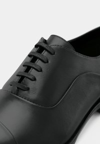 Μαύρο δερμάτινο παπούτσι με λεία υφή, που διαθέτει στρογγυλεμένη μύτη και έξι οπές για κορδόνια. Ο σχεδιασμός περιλαμβάνει διακριτικές ραφές και χαμηλό τακούνι.